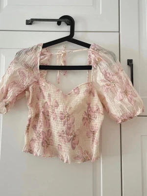 Blommig croppad topp med puffärm - Söt croppad topp med ljusbeige bas och rosa blommigt mönster. Toppen har puffiga korta ärmar, omlott-detalj med knyt i ryggen och en v-ringad front. Materialet är lätt och smockat, perfekt för sommarens varma dagar.