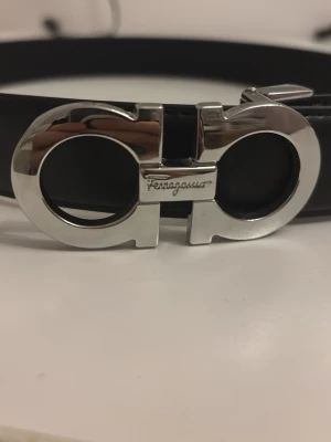 Svart skärp av äkta läder - Snyggt svart skärp av äkta löder med ikoniskt silverfärgat spänne i metall. Skärpet är stilrent och har en klassisk design som passar till många olika outfits. Tillverkat i skinn för en exklusiv känsla. TILLKOMMER MED HÅLGÖRARE!