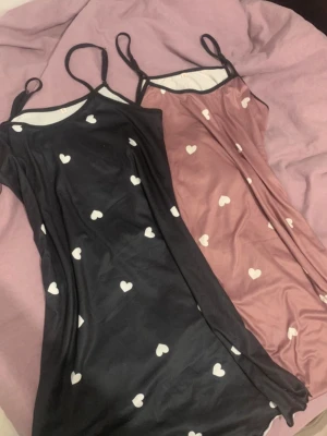 Två nattlinnen med hjärtmönster - Säljer två nattlinnen i mjukt, stretchigt material. Ett är svart och det andra är rosa, båda med vita hjärtan och tunna axelband. Perfekta för mysiga kvällar hemma. Materialet känns svalt och följsamt mot huden.