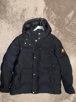Svart dunjacka från Moose Knuckles - Snygg svart dunjacka från Moose Knuckles med huva och guldfärgade detaljer. Jackan har fyra stora fickor framtill, dragkedja och tryckknappar samt logotyp på ärmen. Perfekt för kalla vinterdagar tack vare det tjocka dunfodret. Jackan är knappt använt, och kvittot finns med från johnells. 