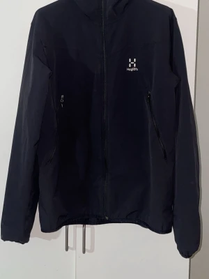 Haglöfs - Snygg svart vindjacka från Haglöfs i storlek XL. Jackan har hel dragkedja framtill, två snedställda fickor med dragkedja och justerbar nederkant. Tillverkad i lätt och slitstarkt material, perfekt för blåsiga dagar.