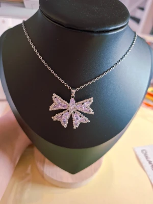 Silverhalsband med glittrande rosett - Snyggt halsband i silverfärg med en stor, glittrande rosett som hänge. Rosetten är täckt av små gnistrande stenar som ger ett lyxigt intryck. Kedjan är tunn och enkel vilket gör att hänget verkligen står ut. Perfekt accessoar för att lyfta din outfit.