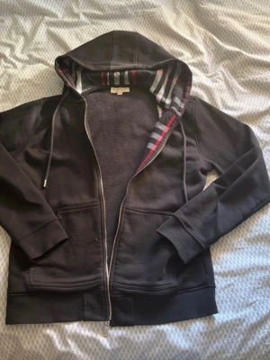 Svart hoodie med dragkedja Burberry - Säljer en svart hoodie från Burberry med dragkedja och klassiskt rutigt foder i huvan och längs insidan. Hoodien har fickor framtill, ribbade muddar och snören i huvan. Materialet är mjukt och bekvämt, perfekt för en chill look.