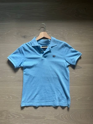 Blå pikétröja från Lexington - Säljer en klassisk blå pikétröja från Lexington med krage och knappar framtill. Tröjan har korta ärmar och ett litet Lexington-emblem på bröstet. Perfekt för dig som gillar en enkel och stilren look.