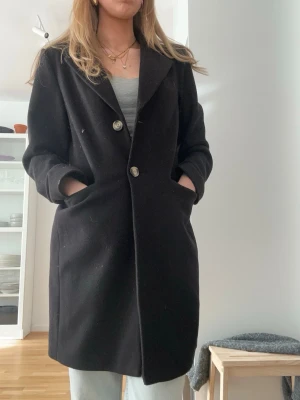 Svart lång kappa med knappar - Stilren svart kappa med två stora knappar framtill och djupa fickor. Klassisk rak passform och enkel krage, perfekt för kyliga dagar. Passar till många olika outfits och ger en snygg siluett. Passar bra i storleken och nästintill oanvänd, säljs pga av att den inte används.