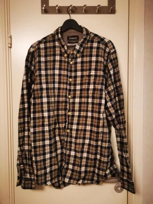 Rutig långärmad skjorta från Sebago - Snygg rutig skjorta från Sebago i svart, vit och beige. Skjortan har klassisk krage, knäppning framtill och långa ärmar. Perfekt för dig som gillar en avslappnad men ändå stilren look.