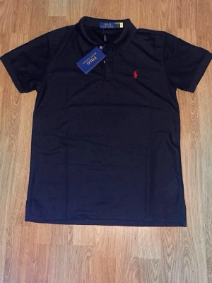 Svart piké från Polo Ralph Lauren - Klassisk svart pikétröja från Polo Ralph Lauren med korta ärmar och röd broderad logga på bröstet. Tröjan har krage och knappar vid halsen. Perfekt för en clean och stilren look.