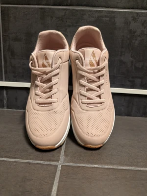 Beige sneakers från Skechers - Snygga beige sneakers från Skechers med perforerad ovandel och klassisk snörning. Skorna har en vit sula med bruna detaljer och logotyp i guld på plösen. Perfekt för dig som vill ha en stilren och bekväm sneaker till vardags. Storlek 37