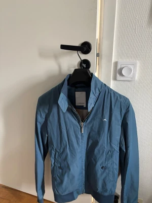 Blå vindjacka från J.Lindeberg - Snygg blå vindjacka från J.Lindeberg med dragkedja framtill och klassisk krage. Jackan har två fickor med knappstängning och diskret logotyp på bröstet. Materialet är lätt och passar perfekt för blåsiga dagar.
