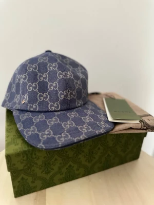 Blå GG-monogram keps från Gucci - Snygg blå keps Gucci i denim-look med det klassiska GG-monogrammet över hela kepsen. Justerbar rem baktill för perfekt passform. Kepsen har böjd skärm och är tillverkad i slitstarkt tyg. Perfekt accessoar för dig som vill sticka ut med lyxig streetstyle.