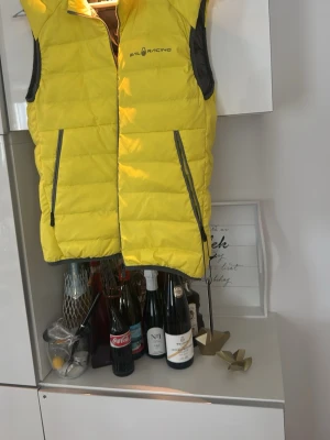 Gul dunväst från Sail Racing XS - Cool gul dunväst från Sail Racing med svarta detaljer och två dragkedjeförsedda fickor. Västen har hög krage och är perfekt för lager-på-lager. Materialet är quiltat och ger en sportig look.