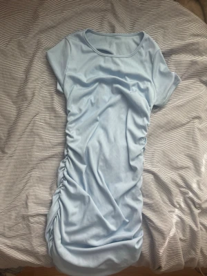 Ljusblå ribbad kortklänning - Säljer en ljusblå ribbad kortklänning med snörning på sidan. Klänningen har rund halsringning och korta ärmar, perfekt för dig som gillar en enkel men snygg look. Materialet är mjukt och stretchigt, vilket gör den bekväm att bära.