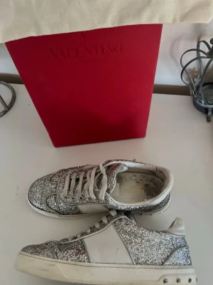 Valentino glitter sneakers silver - Säljer ett par riktigt snygga Valentino sneakers med silverglitter och vita detaljer. Skorna har klassisk låg profil, vita snörningar och coola nitar på hälen. Perfekt för dig som vill sticka ut och addera lite bling till din outfit.