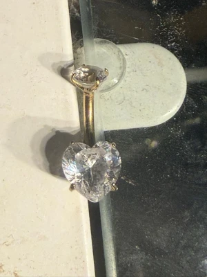 Guldpläterad navelpiercing med hjärtformad sten - Snygg navelpiercing med en stor, hjärtformad klar sten och en mindre rund sten upptill. Staven är guldpläterad och ger ett lyxigt intryck. Perfekt för dig som vill ha något extra och glittrigt till din look.köpt i Spanien för 200. råstfri. Aldirg använd då mitt hål redan hade växt igen innan jag köpte.