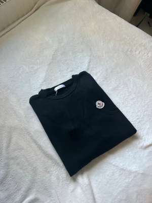 Svart Moncler tröja med logotyp - Snygg svart tröja från Moncler med rund hals och lång ärm. Tröjan har Monclers ikoniska logotyp broderad på bröstet. Materialet är mjukt och bekvämt, perfekt för dig som vill ha en stilren look med exklusiv känsla.