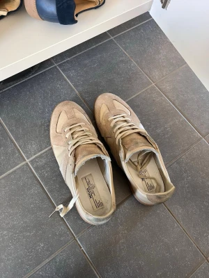 Maison Margiela gats - Säljer ett par beige sneakers från Maison Margiela, tillverkade i Italien. Skorna har en klassisk låg modell med både mocka och läderdetaljer, samt vita snören. Perfekt för dig som gillar stilrena och exklusiva sneakers. Köptes secondhand 
