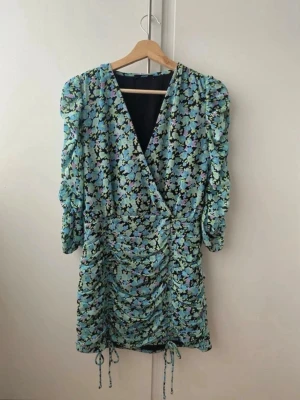 Blommig klänning  - Snygg blommig klänning i blått med trekvartsärm och v-ringning. Klänningen har en kort längd och dragsko-detaljer på sidorna för en justerbar passform. Perfekt för sommarens alla tillfällen! 