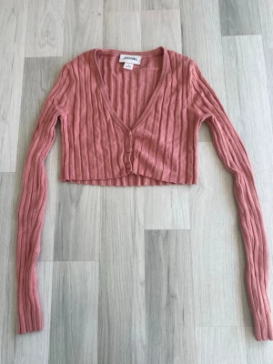 Rosa ribbad cropped kofta Monki - Säljer en rosa ribbad cropped kofta från Monki med djup v-ringning och tre knappar framtill. Koftan har långa ärmar och är tillverkad i ett mjukt stickat material som ger en skön känsla. Finns lite tecken på användning men inget som syns. Storlek s passar även som xs 