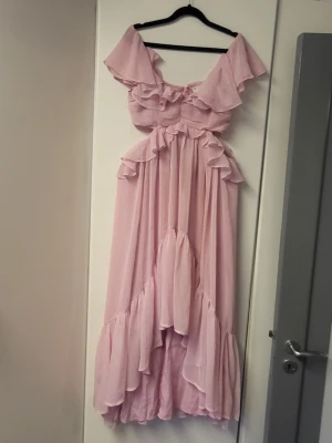 Ljusrosa långklänning med volanger - Romantisk ljusrosa långklänning med volanger och offshoulder-design. Klänningen har omlott-detaljer och är tillverkad i ett lätt och flödande material som ger en drömmig känsla. Perfekt för dig som vill sticka ut med en feminin och elegant look.