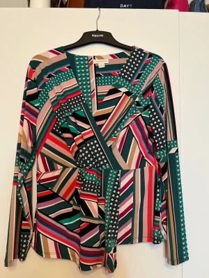 Färgglad mönstrad blus från Monsoon - Säljer en långärmad blus från Monsoon med ett coolt geometriskt mönster i grönt, rosa, svart, beige och rött. Blusen har en v-ringning och är gjord i ett mjukt, lätt material som känns skönt mot huden. Perfekt för dig som gillar färg och unika prints.