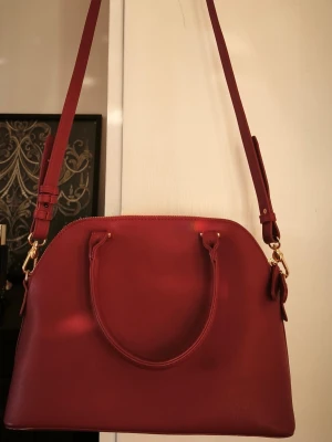 Mörkröd handväska med axelrem Misako - Mörkröd (den är mörkare än på bild, nästan lite åt lila) handväska med guldfärgade detaljer. Väskan har ett huvudfack som stängs med dragkedja. Inuti finns ett mindre sidofack med dragkedja. Väskan har två handtag och en avtagbar, justerbar axelrem.   Använd vid ett tillfälle. En söm är ljusare längst ner på en sida, var så här redan vid köpet.