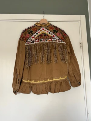 Brun boho-jacka med brodyr och fransar - Unik brun jacka i boho-stil med färgglad brodyr och långa fransar på ryggen. Den har volangkant nedtill och detaljer i gult samt mönstrade axelpartier. 