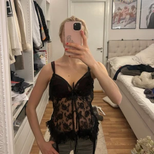 Svart spetsbody med rosett - Säljer en svart topp i transparent spets med dekorativ rosett framtill. Den har tunna axelband och vacker spetsdetalj över hela plagget. Perfekt för dig som gillar en romantisk och edgy stil.