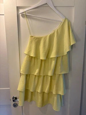 Gul omlottklänning med volanger från Zara - Säljer en ljusgul kortklänning från Zara med flera lager volanger och oneshoulder-design. Klänningen har ett axelband och är perfekt för sommarens alla tillfällen. Materialet är mjukt och lätt, troligen bomull eller syntet.