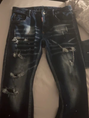 Dsquared2  - Säljer ett par blå jeans från Dsquared2 med slitna detaljer och trasiga partier på framsidan. Jeansen har en cool tvätt med ljusa partier och klassisk femficksdesign. Materialet är denim och passformen är rak. Perfekt för dig som gillar en edgy streetstil.