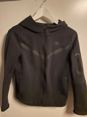 Svart Nike hoodie med dragkedja - Cool svart hoodie från Nike med dragkedja och huva. Diskret Nike-logga på bröstet och snygga detaljer på ärmarna, inklusive en liten ficka med dragkedja på ena armen. Perfekt för dig som gillar sportig stil och komfort.