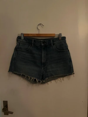 Blå denim shorts från Wrangler - Säljer ett par klassiska blå denim shorts från Wrangler med hög midja och råa, fransiga benslut. Shortsens femficksdesign och tydliga bakfickor med W-stitch.  