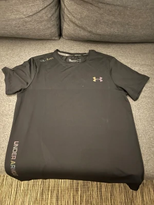 Svart tränings-tshirt Under Armour - Cool svart tränings-tshirt från Under Armour med logga i regnbågsfärger på bröstet och text på axeln samt längs sidan. Materialet är lätt och snabbtorkande, perfekt för gym eller löpning. Snygg passform och modern look.
