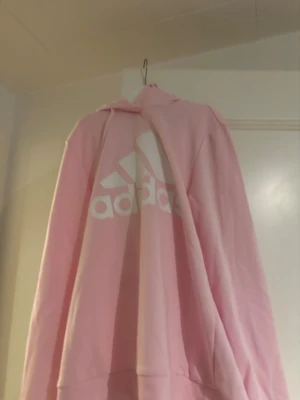 Rosa hoodie från Adidas med logga - Säljer en ljusrosa hoodie från Adidas med stor vit logga på bröstet. Hoodien har en klassisk huva och ribbade muddar vid ärmslut och nederkant. Materialet är mjukt och bekvämt, perfekt för en avslappnad stil.