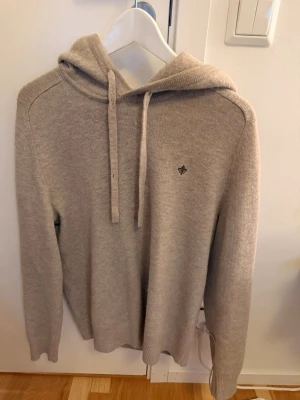 Beige stickad hoodie med huva - Mysig beige stickad hoodie med huva och dragsko. Tröjan har ett diskret broderat emblem på bröstet och är tillverkad i ett mjukt material som känns skönt mot huden. Perfekt för dig som gillar en clean och stilren look. Pris kan diskuteras. Nypris 2000kr skicka gärna bud. Passar också