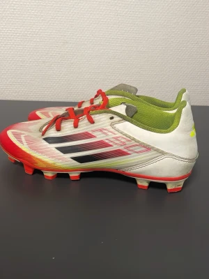 Adidas f50 - Säljer ett par snygga fotbollsskor från Adidas i vitt med röda och svarta detaljer samt limegröna insidor. Skorna har klassiska dobbar för gräsplan och är tillverkade i syntetmaterial. Perfekta för dig som vill sticka ut på planen!