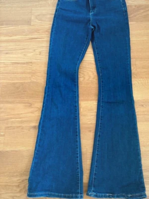 Blå bootcut jeans med hög midja - Säljer ett par blå bootcut jeans med hög midja och klassisk femficksdesign. Jeansen har en snygg utsvängd passform som ger en retrovibe och är tillverkade i denim. Perfekta för dig som gillar en trendig och avslappnad look.