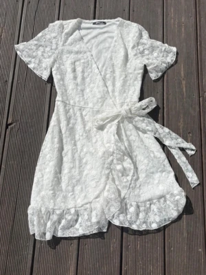 Vit spetsjumpsuit med volang - Supersöt vit jumpsuit i spets med korta ärmar och omlottmodell. Har knytband i midjan och volangkant nedtill för extra detalj. Perfekt för sommarens alla tillfällen! Materialet är genombruten spets med blommönster.