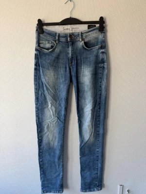Sams Jeans - Ett par blå slim fit jeans från Sams Jeans med klassisk femficksdesign och slitningar på framsidan. Jeansen har en knapp och dragkedja framtill samt läderpatch bak. Materialet är denim och passformen är smal.