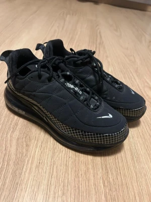 Nike Air Max 720-818 svarta sneakers - Svarta Nike Air Max 720-818 sneakers med quiltad ovandel, vita detaljer och reflekterande logga. Skorna har en hög sula med rutmönster längs kanten och synlig Air Max-dämpning. Snygg futuristisk look med snörning och dragflik bak.