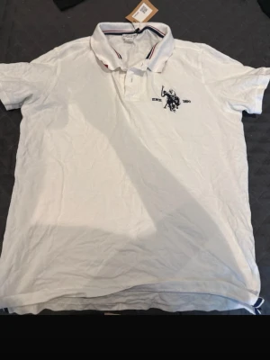 Vit pikétröja från U.S. Polo Assn - Klassisk vit pikétröja från U.S. Polo Assn med broderad logga på bröstet. Tröjan har krage med knappslå och korta ärmar. Perfekt till en avslappnad men ändå stilren look.