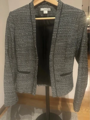 Svart och vit kavaj från H&M - Stilren svart och vit kavaj från H&M med diskret mönster och raka linjer. Kavajen har två dekorativa fickor framtill och är tillverkad i ett strukturerat tyg som ger en modern look. Perfekt för dig som gillar klassiska detaljer och enkel design.