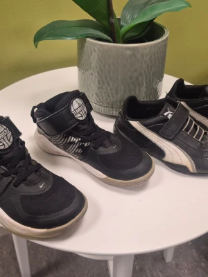 2-pack Nike Team Hustle D9, Svarta Puma Storlek 28 - Två par svarta sneakers, ett från Nike med snörning och kardborreband, och ett från Puma med kardborreband och vit detalj på sidan. Båda har sportig design och är tillverkade i syntetmaterial med mesh och skinnliknande ytor. Perfekta för dig som gillar klassiska streetstyles.