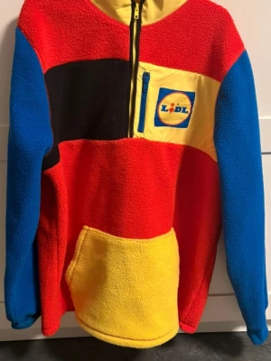 Färgglad stickad fleece från Lidl - Säljer en unik och färgglad stickad fleece från Lidl med halv dragkedja. Tröjan har blå ärmar, röd kropp, svart och gul panel samt en gul ficka framtill. Lidl-loggan finns på bröstet. Perfekt för dig som gillar att sticka ut och vill ha något riktigt coolt.
