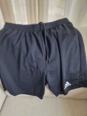Svarta träningsshorts från Adidas - Säljer ett par svarta träningsshorts från Adidas med klassisk logga på benet. De har elastisk midja med snörning och är tillverkade i ett lätt syntetmaterial som passar perfekt för sport och aktivitet. Sköna och funktionella shorts med normal passform. Vet ej storlek.  Möjligen M