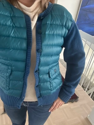 Blå quiltad cardigan - Snygg blå cardiganjacka med quiltad, vadderad front och stickade ärmar samt rygg. Två stora fickor framtill med lock och knappar. Ribbstickade muddar och knappstängning hela vägen. Perfekt för lager på lager och chill stil.