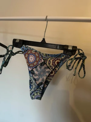 Färgglada bikiniunderdelar med knyt - Snygga bikiniunderdelar med färgglatt, bohemiskt mönster i blått, grönt och beige. Modellen har knyt i sidorna för justerbar passform och är perfekt för strandhäng. Materialet känns mjukt och följsamt mot huden.