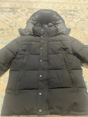 Svart pufferjacka från Canada Goose - Säljer en svart pufferjacka från Canada Goose med hög krage, stor huva och diskret logotyp på ärmen. Jackan har tryckknappar framtill och två stora fickor. Perfekt för kalla dagar och riktigt snygg streetstil.