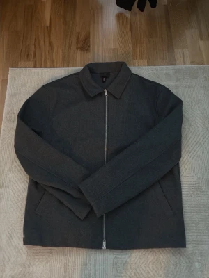 Mörkgrå overshirt med dragkedja - Stilren mörkgrå overshirt från H&M med rak passform och klassisk krage. Jackan har en silverfärgad dragkedja framtill och två snedställda fickor. Materialet ser ut att vara en mjuk ullblandning som ger en clean look.