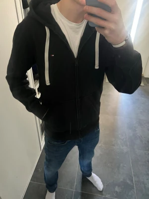 Zip hoodie med vita snören  - Säljer en svart hoodie från Tommy Hilfiger med dragkedja och vita snören. Klassisk liten logga på bröstet och stor huva. Perfekt till jeans eller joggers för en chill look. Mjuk bomullskänsla och fickor framtill.