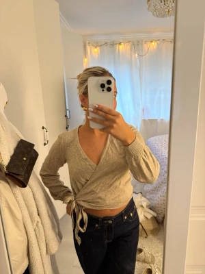 Beige omlottstickad tröja - Säljer en beige stickad omlottopp med långa ärmar och knytband i midjan. Tröjan har en djup v-ringning och ribbade muddar vid ärmsluten. Perfekt att styla med jeans eller kjol för en trendig look.
